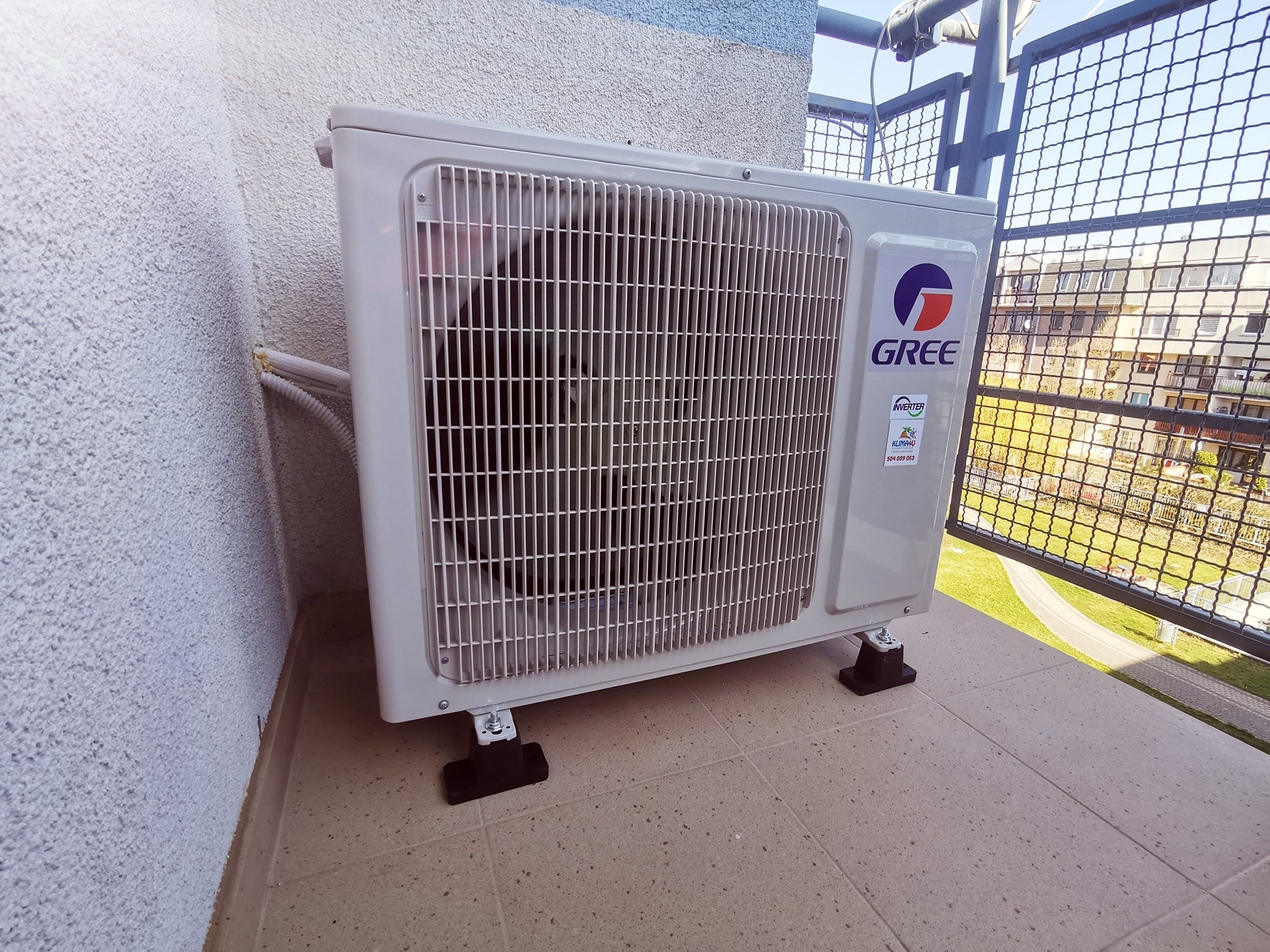 Zewnętrzna jednostka klimatyzacji Gree zamontowana na balkonie z widokiem na osiedle mieszkaniowe, widoczne przewody przy ścianie i regulowane nóżki montażowe.
