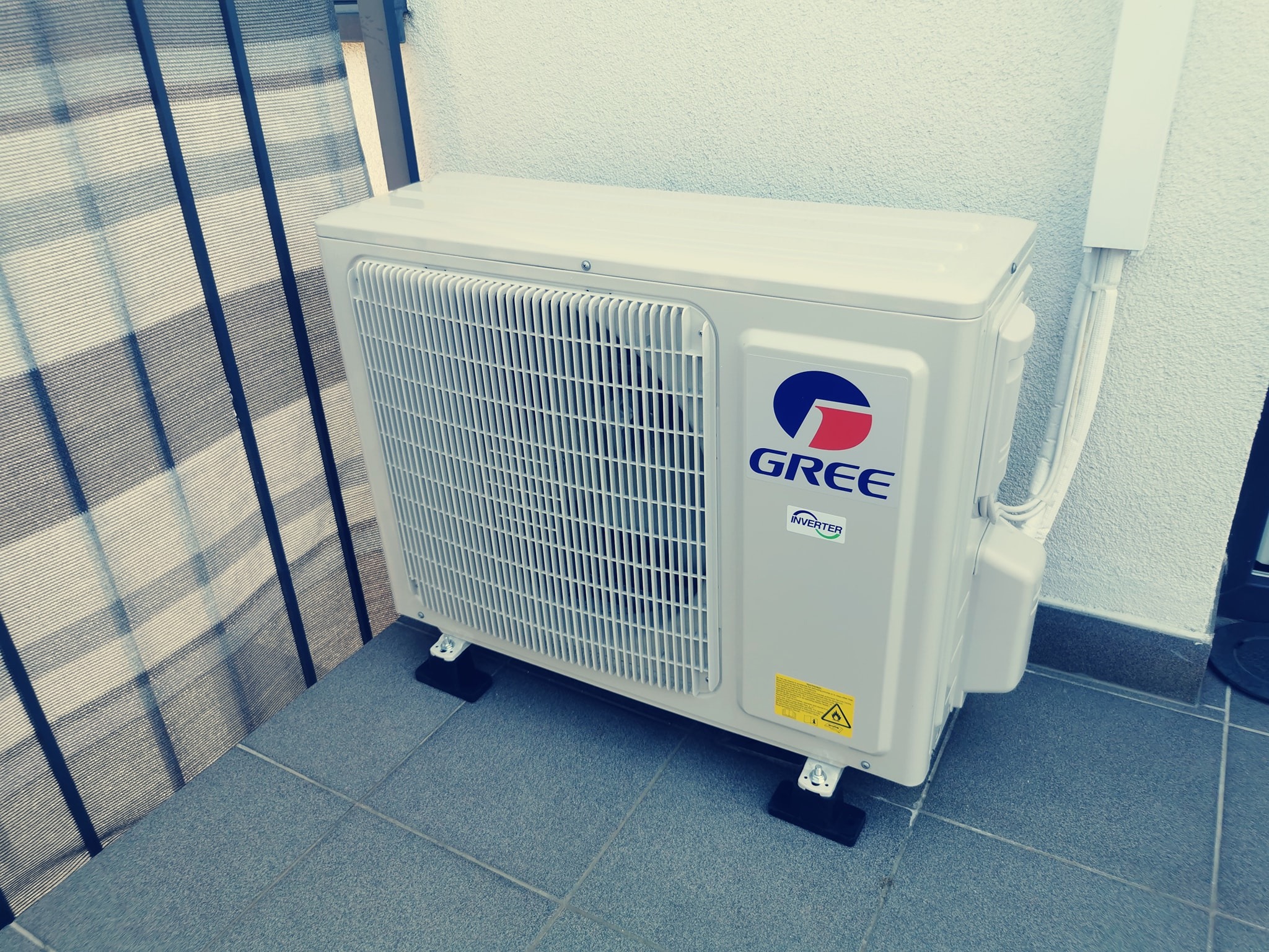 Biała zewnętrzna jednostka klimatyzacji marki Gree zamontowana na balkonie, widoczne przewody zabezpieczone listwą, ściana o fakturze baranka i szara podłoga z płytek.