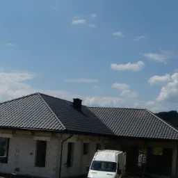 Dom w trakcie budowy z nowym, ciemnoszarym dachem z dachówki ceramicznej; biały samochód dostawczy zaparkowany przed budynkiem; w tle zalesione wzgórza i błękitne niebo z chmurami.