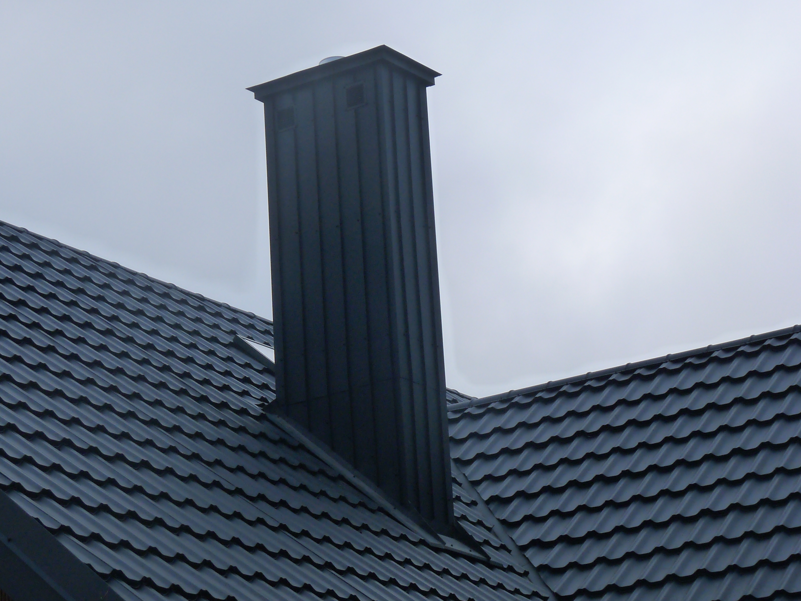 Nowoczesny, ciemny dach z blachy trapezowej z kominem o kwadratowym przekroju na tle pochmurnego nieba.