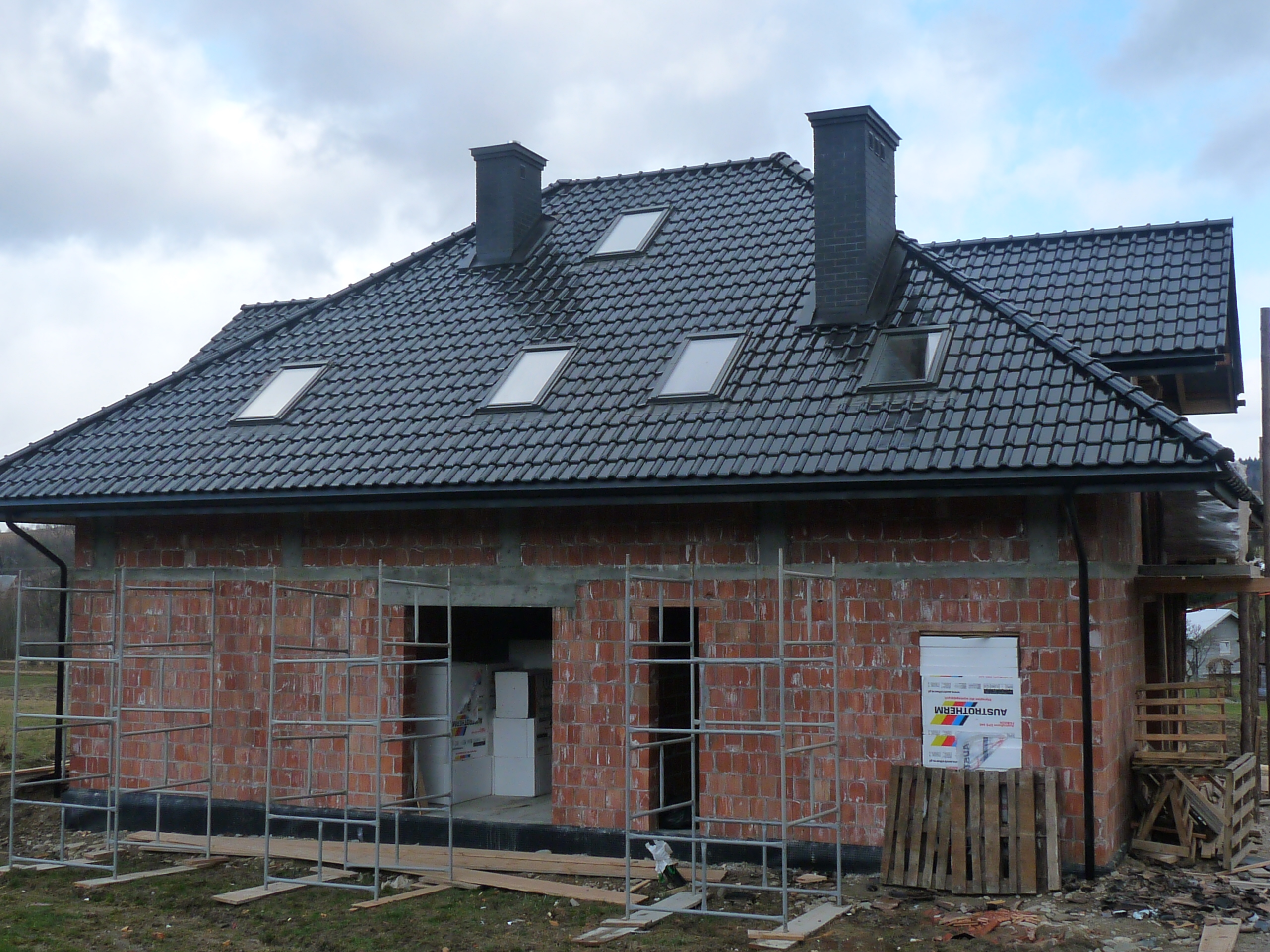 Dom w trakcie budowy z widoczną konstrukcją dachu pokrytą czarną dachówką i oknami dachowymi, murowane kominy, ściany z czerwonej cegły w trakcie wznoszenia, rusztowania wokół budynku, materiały...