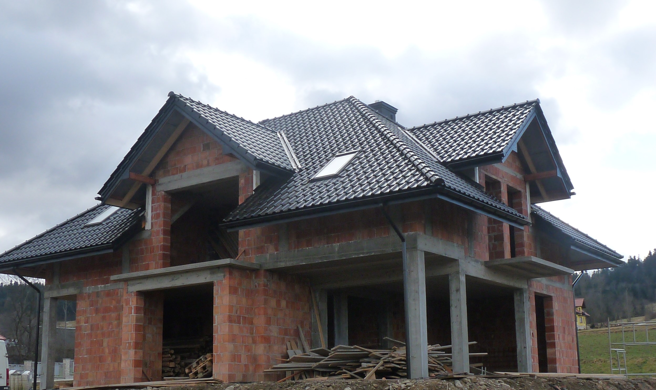 Surowy budynek w trakcie budowy z ciemną dachówką, widoczne elementy więźby dachowej i częściowo murowane ściany z czerwonej cegły, na tle pochmurnego nieba i zalesionego wzgórza.
