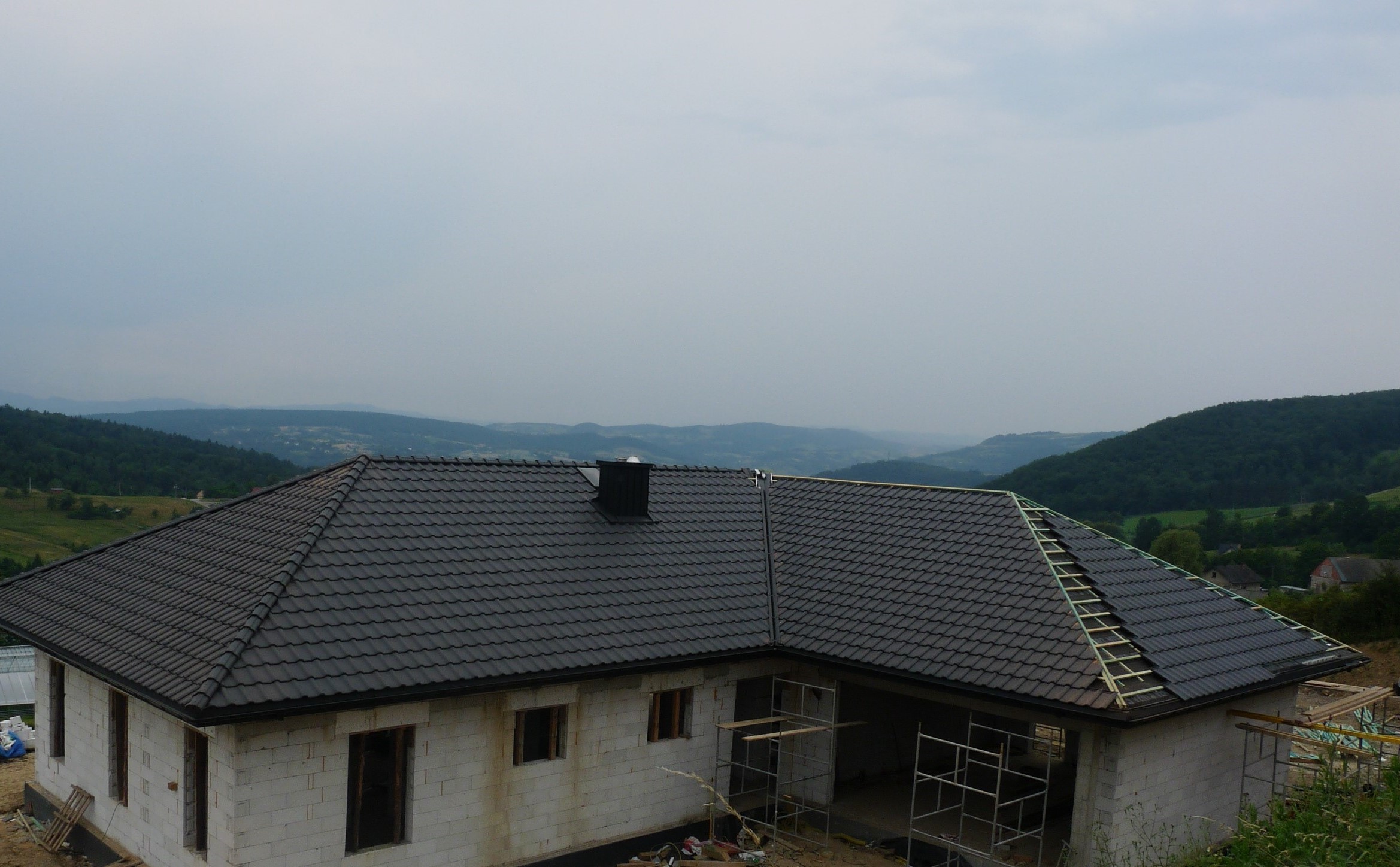 Dom w trakcie budowy z ciemnoszarą dachówką na połaci dachu, częściowo wykończoną, widoczne rusztowania i krajobraz pagórkowaty w tle.