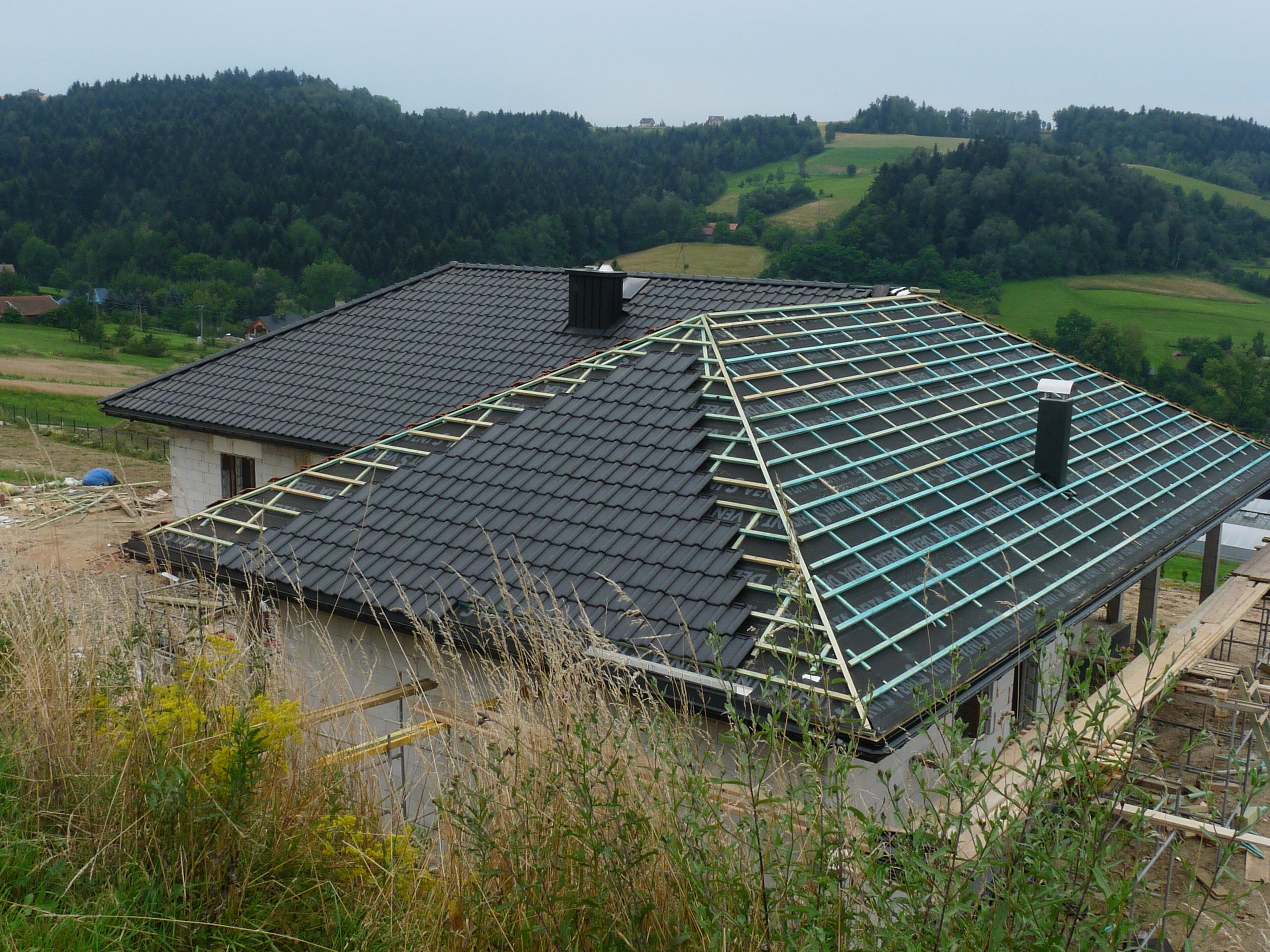 Dom w trakcie wymiany dachu z widoczną częścią dachu pokrytą czarną dachówką i odsłoniętą więźbą dachową, na tle pagórkowatego krajobrazu.