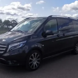 Czarny, luksusowy van Mercedes Vito z relingami dachowymi, zaparkowany na asfalcie z widokiem na pole i pochmurne niebo.