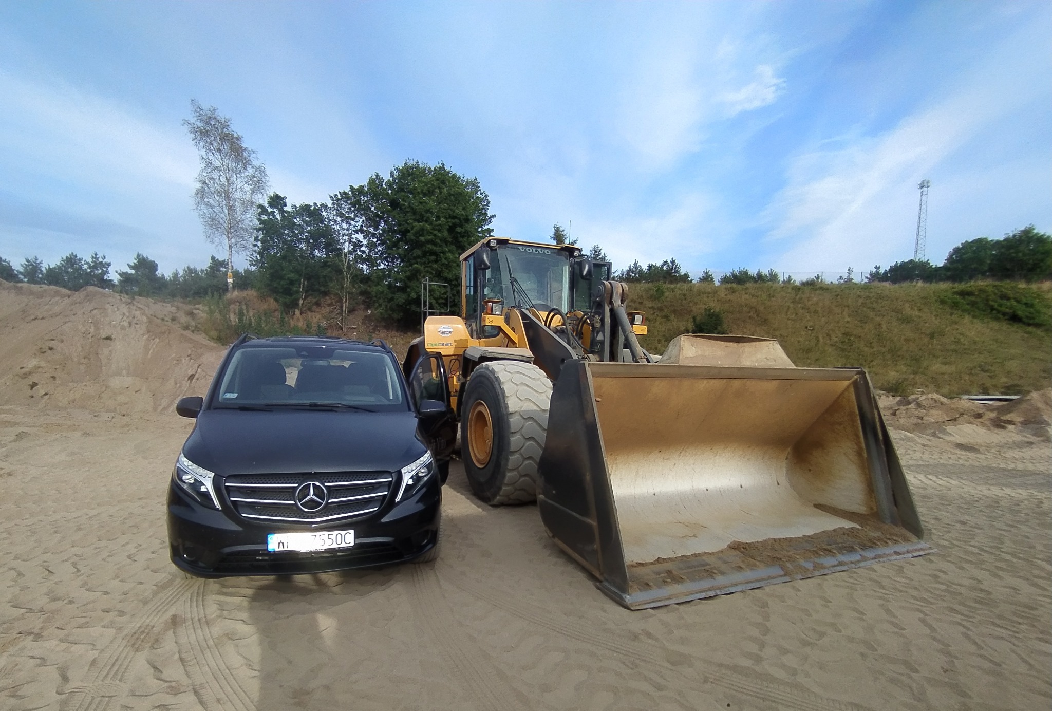 Czarny bus Mercedes i żółta ładowarka Volvo na piaszczystym terenie z pagórkami i drzewami w tle, błękitne niebo z chmurami.