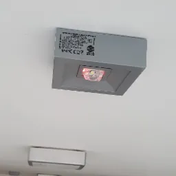 Szary, kwadratowy moduł oświetlenia awaryjnego LED, model Lovato N, zamontowany na białym suficie, widoczne parametry techniczne na obudowie.