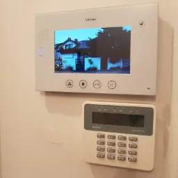 Panel wideodomofonu Vidos z podglądem posesji oraz klawiatura alarmowa Satel Perfecta zamontowane na ścianie wewnątrz budynku, obok dekoracyjnej ramy lustra.