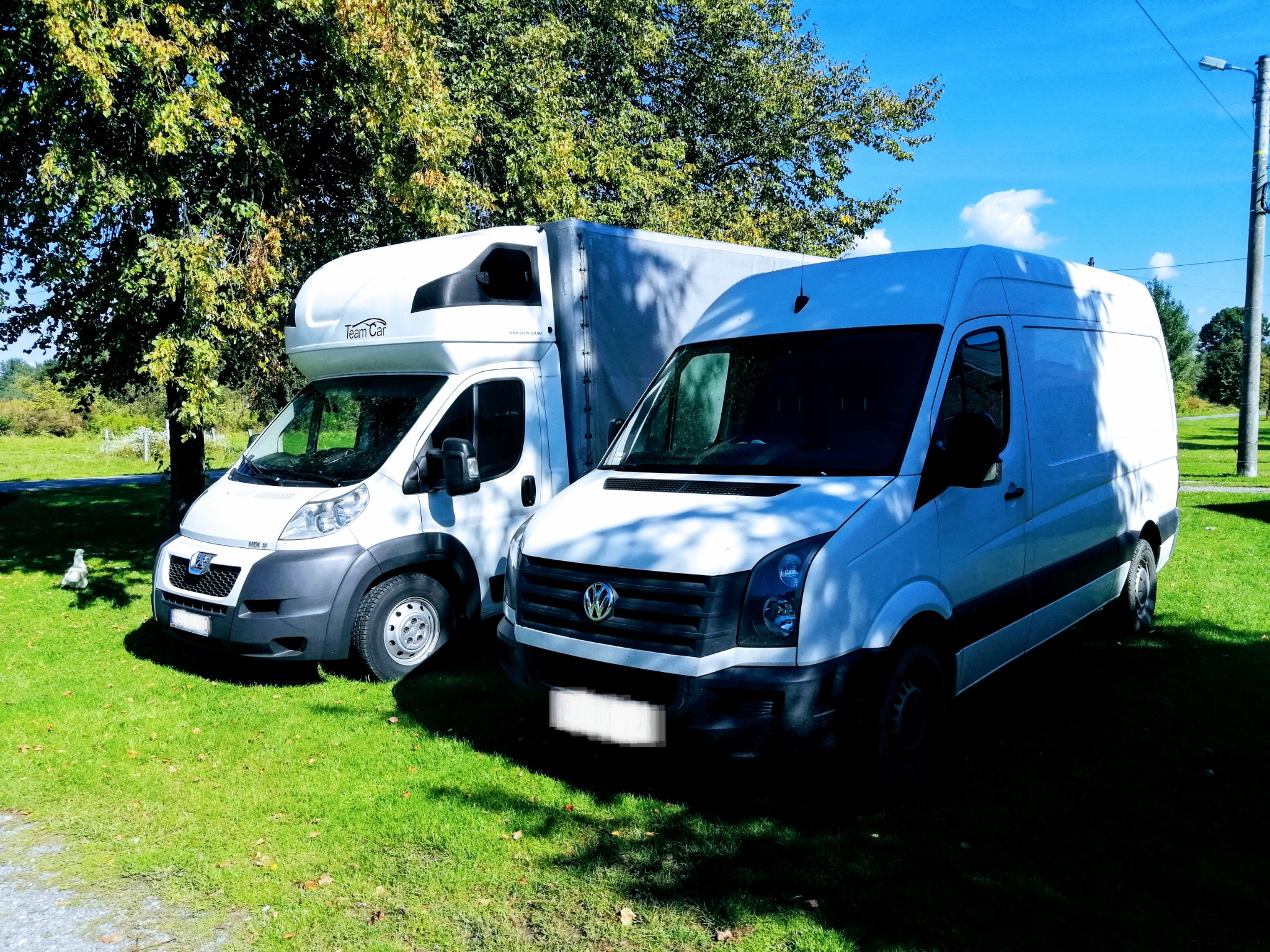 Dwa białe busy, jeden z nadbudówką, zaparkowane na trawniku w słoneczny dzień, widoczne drzewa i niebo.