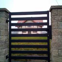 Nowoczesna, pozioma brama wjazdowa z czarnych, płaskich profili, osadzona na kamiennych słupkach, z widocznym domem w tle.