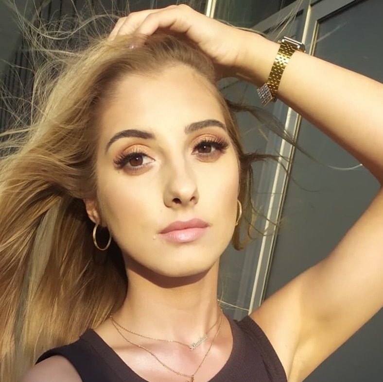 Portret kobiety z delikatnym makijażem oczu i ust, złote kolczyki koła i naszyjniki, włosy rozwiane na wietrze, złoty zegarek na ręce.