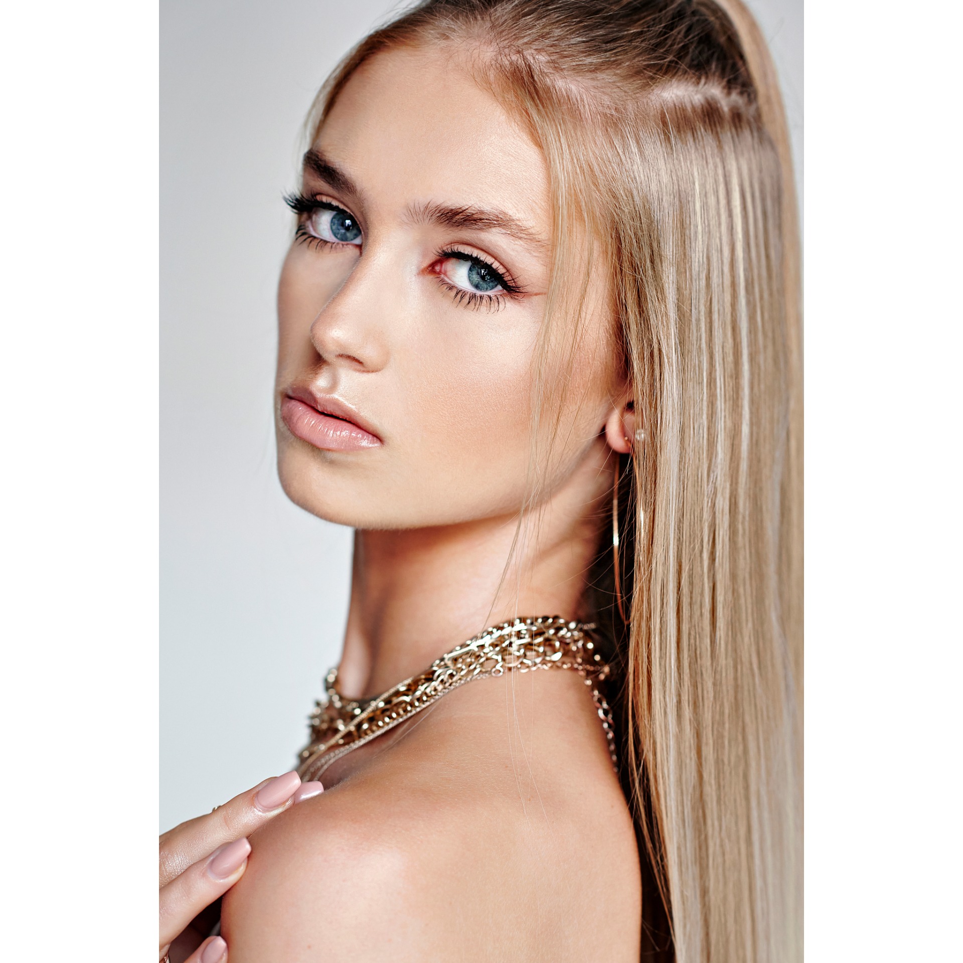 Portret młodej kobiety z blond włosami, subtelnym błyszczącym makijażem, podkreślonymi rzęsami i złotą biżuterią, pozującej na jasnym tle.