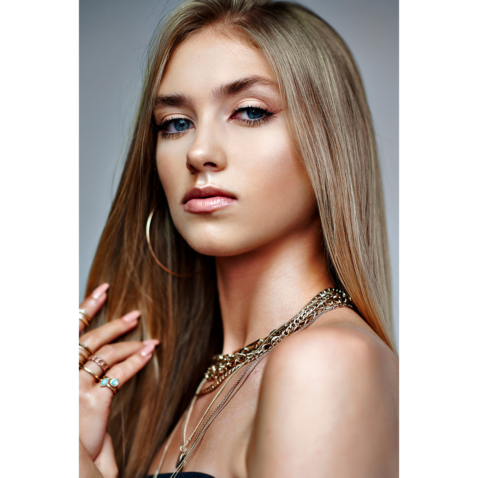 Portret młodej kobiety z blond włosami, subtelnym podkreśleniem oczu i błyszczącymi ustami, ozdobionej złotą biżuterią i pierścionkami z turkusem, na jednolitym szarym tle.