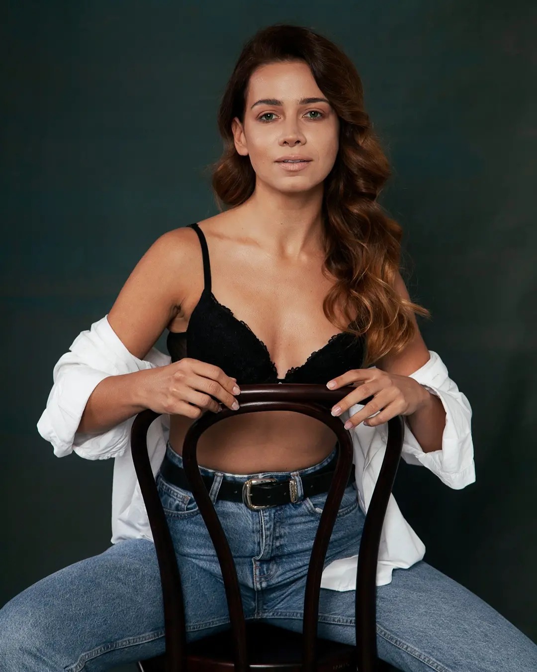 Portret kobiety siedzącej na krześle, z delikatnym makijażem, w czarnym biustonoszu i rozpiętej białej koszuli, na ciemnym tle.