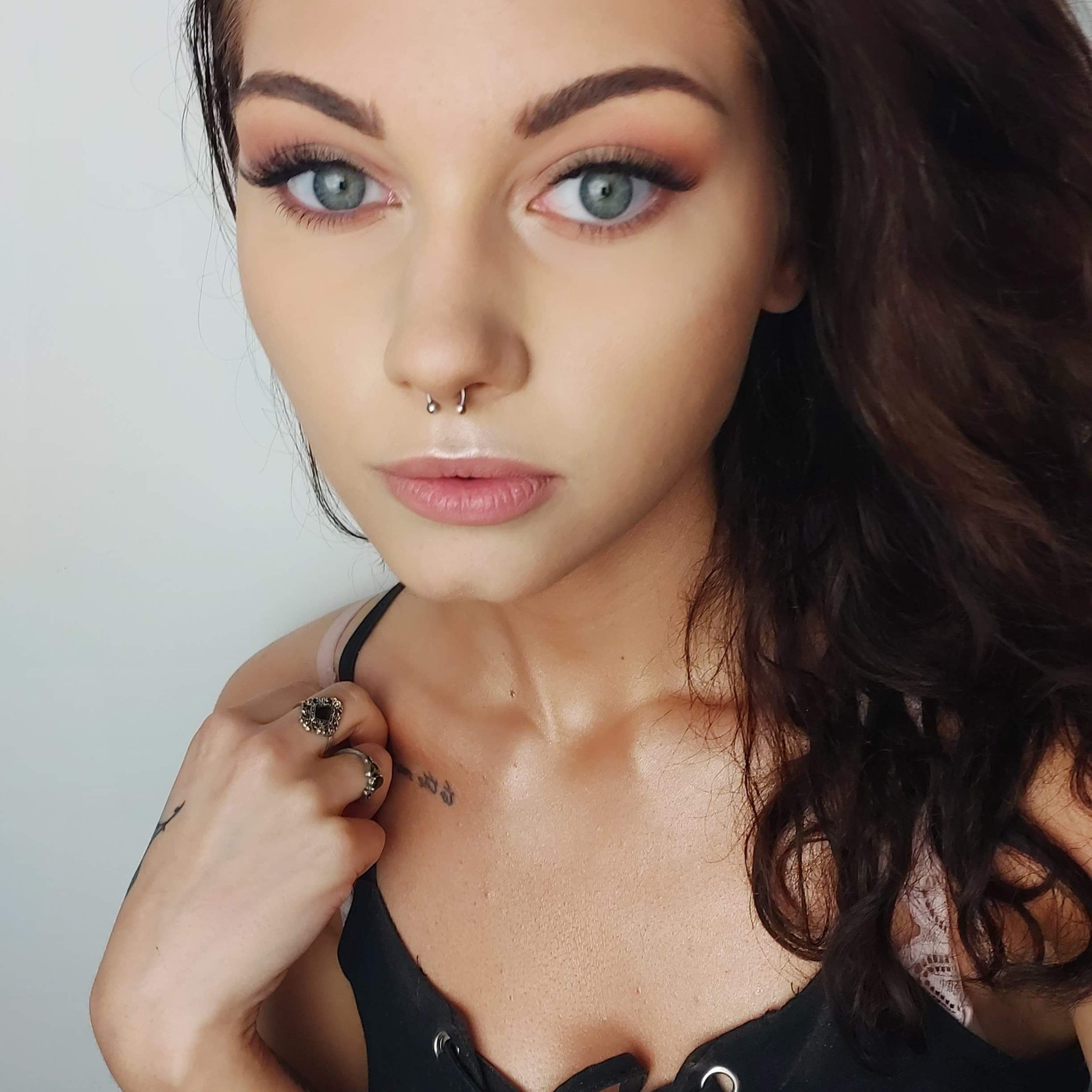 Portret kobiety z delikatnym dziennym makijażem, kolczykiem w nosie typu septum i ciemnymi włosami, w czarnej bluzce na ramiączkach z koronką, patrzącej w obiektyw.