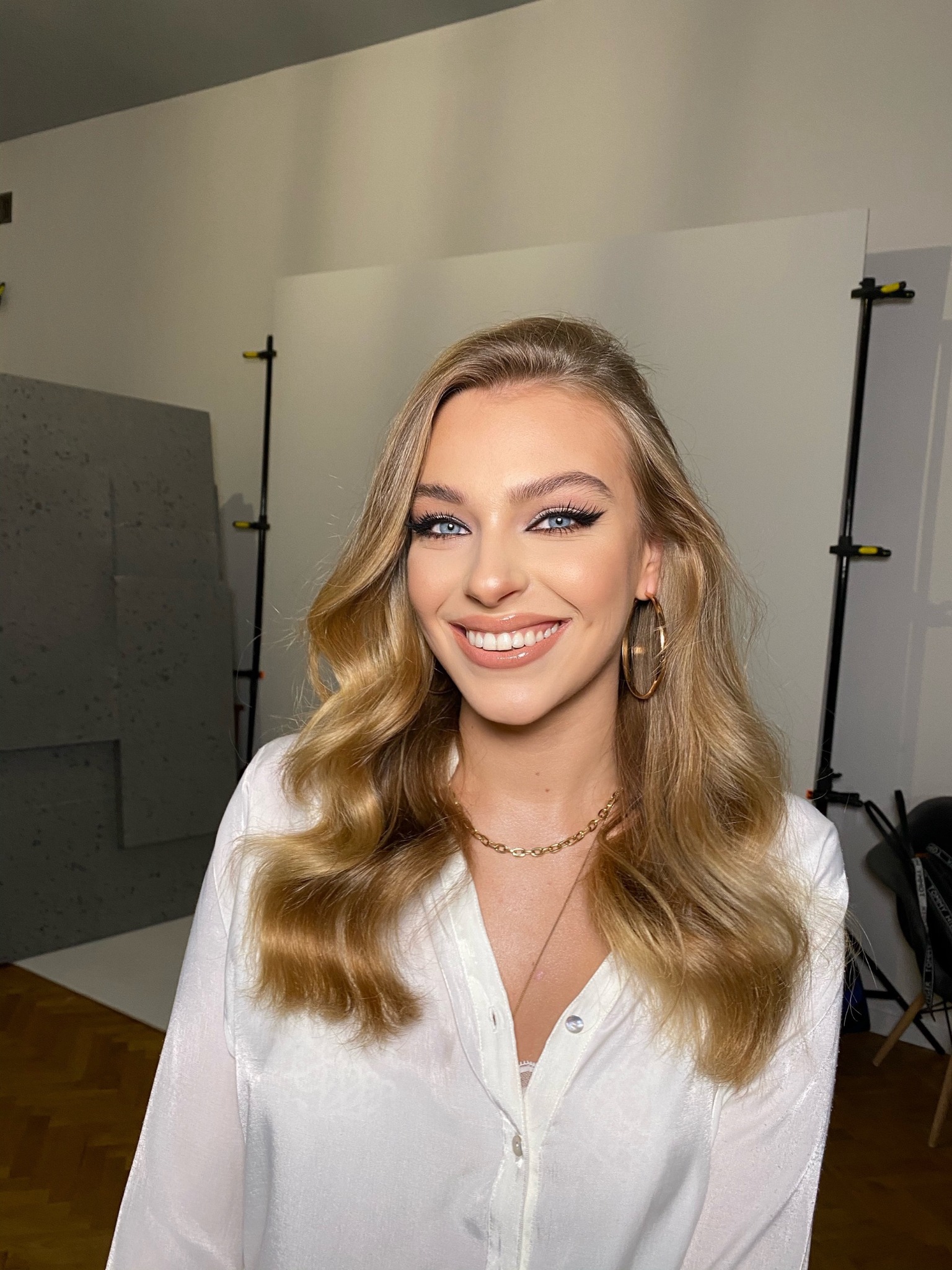 Portret uśmiechniętej blondynki z wyraźnie podkreślonymi oczami czarnym eyelinerem i długimi rzęsami, złote kolczyki koła i łańcuszek na szyi, w tle studio fotograficzne z blendą i statywami...