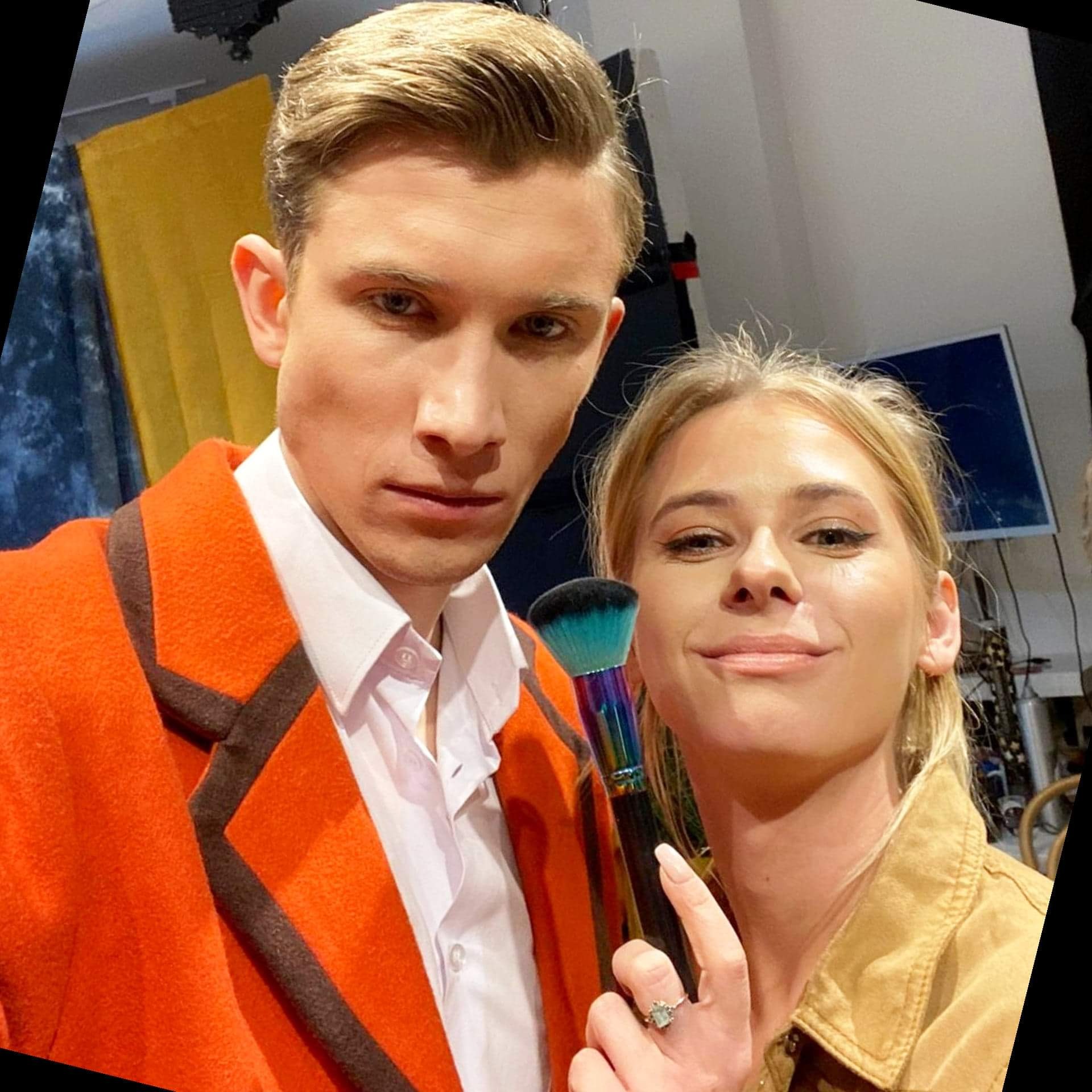 Selfie kobiety trzymającej pędzel do pudru z tęczową rączką, obok mężczyzny w pomarańczowym płaszczu z delikatnym make-upem.
