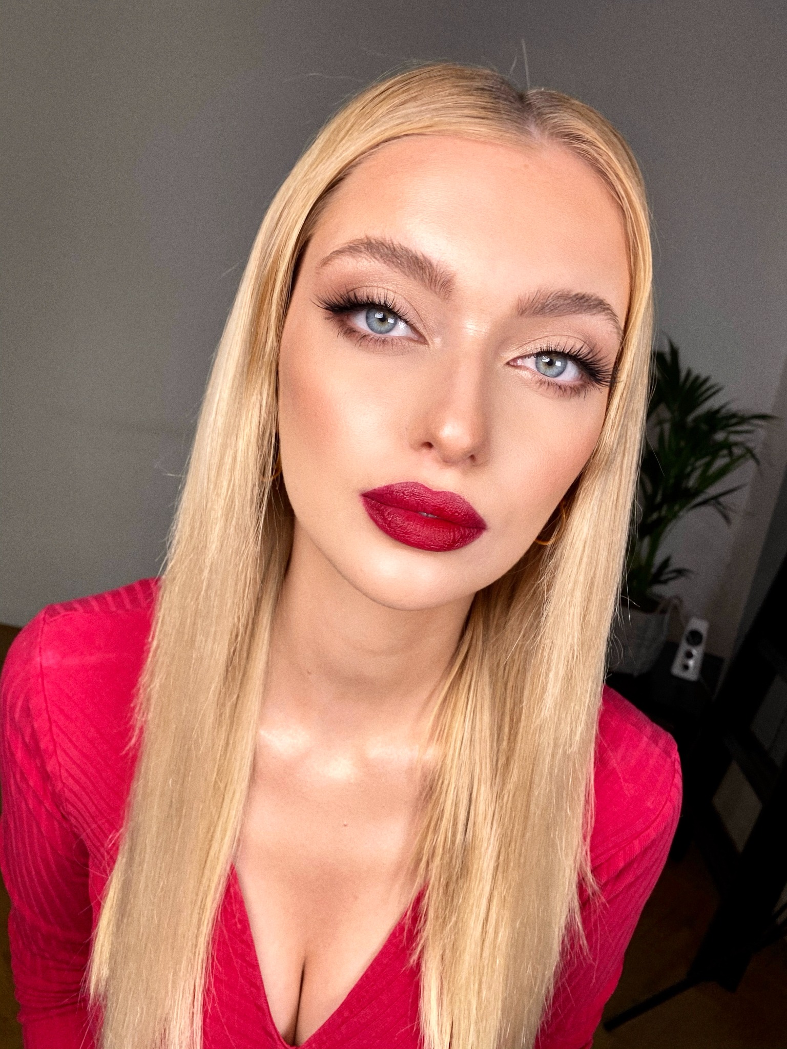 Portret blondynki z wyrazistymi, czerwonymi ustami, podkreślonymi rzęsami i delikatnym makijażem oczu, ubrana w czerwoną bluzkę.