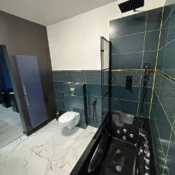 Nowoczesna łazienka z czarną wanną z hydromasażem, toaletą podwieszaną i ciemnoniebieskimi płytkami z geometrycznym wzorem oraz złotymi fugami, biała marmurkowa podłoga.