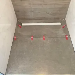 Ułożone płytki podłogowe w kabinie prysznicowej, z widocznym odpływem liniowym i czerwonymi klipsami poziomującymi, ściany wyłożone białymi płytkami imitującymi marmur oraz drewnopodobnymi płytkami.