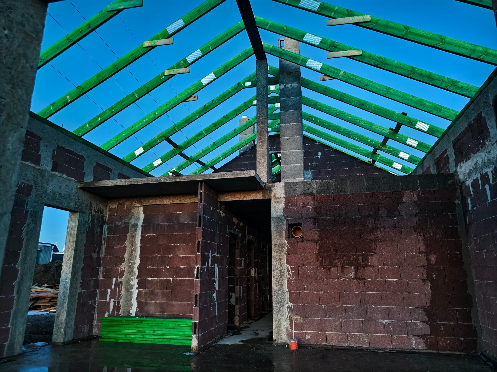 Surowy stan domu w trakcie budowy: czerwona cegła, zielona więźba dachowa na tle błękitnego nieba. Widoczne elementy konstrukcyjne i otwory okienne.