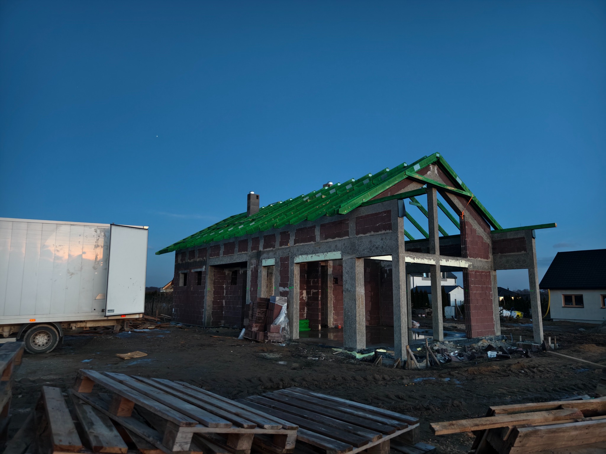 Stan surowy domu jednorodzinnego z cegły, z zieloną konstrukcją dachu, widok z zewnątrz w Oleśnicy. Na pierwszym planie palety budowlane.