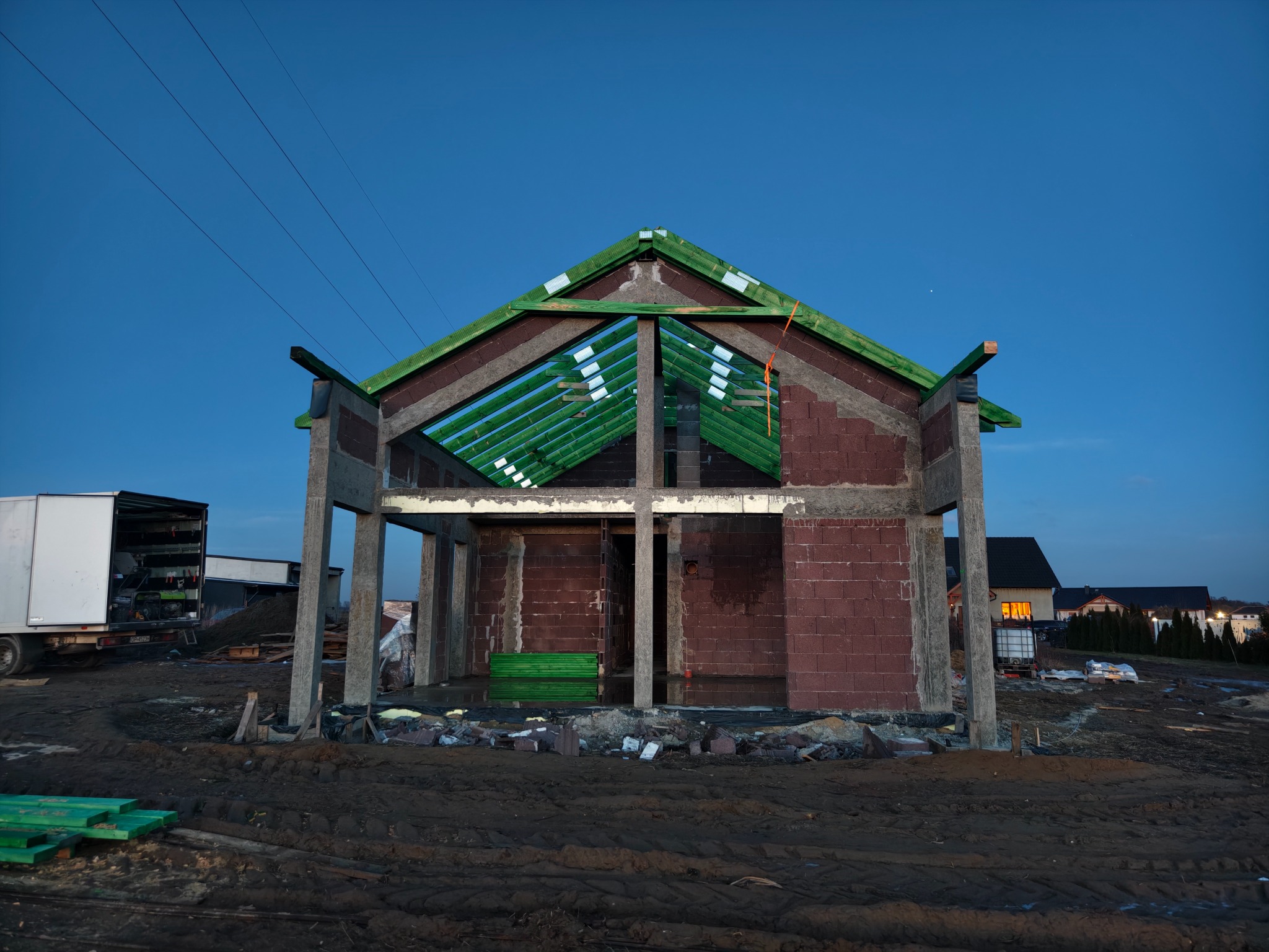 Wznoszenie domu z czerwonej cegły: surowa konstrukcja z zielonymi elementami dachu na tle błękitnego nieba. Widoczny teren budowy i częściowo zaparkowany biały samochód dostawczy.