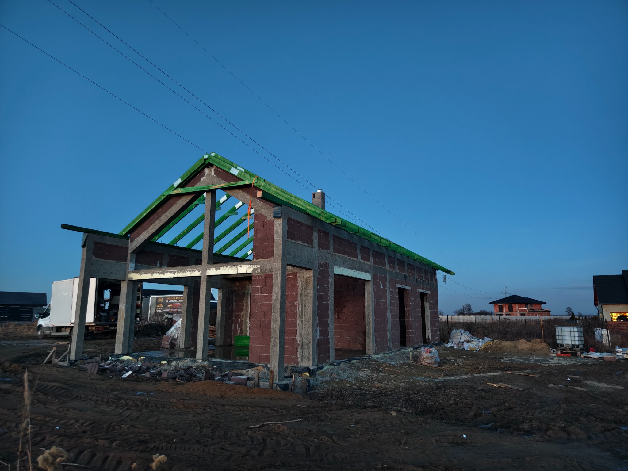Niedokończony dom z czerwonej cegły i betonu w surowym stanie, z widoczną konstrukcją dachu i elementami szalunku, na tle błękitnego nieba o zmierzchu.