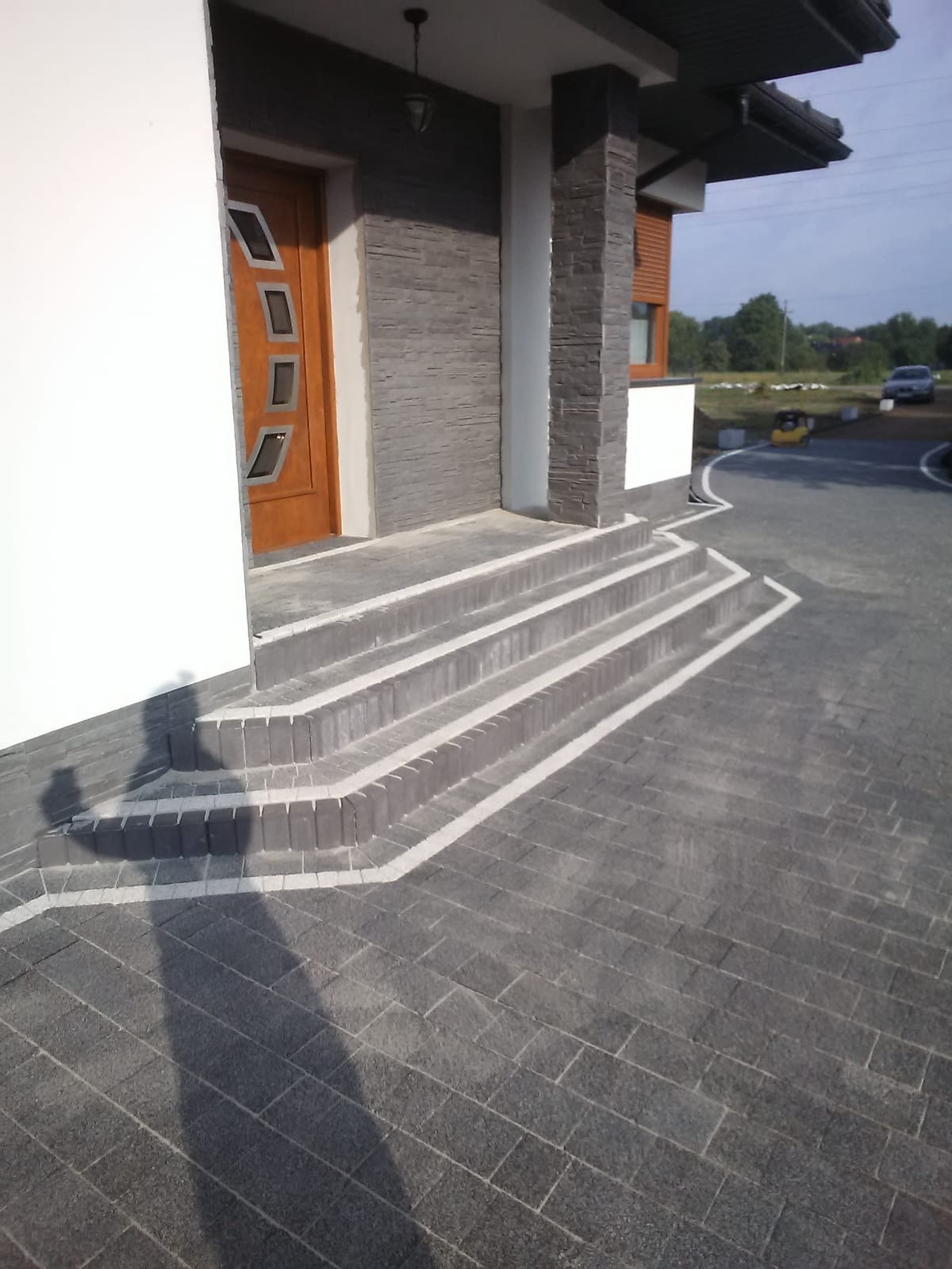 Nowoczesne wejście do domu z kamiennymi schodami, kostką brukową w odcieniach szarości i białymi liniami akcentującymi krawędzie, w tle widoczny budynek i zieleń.