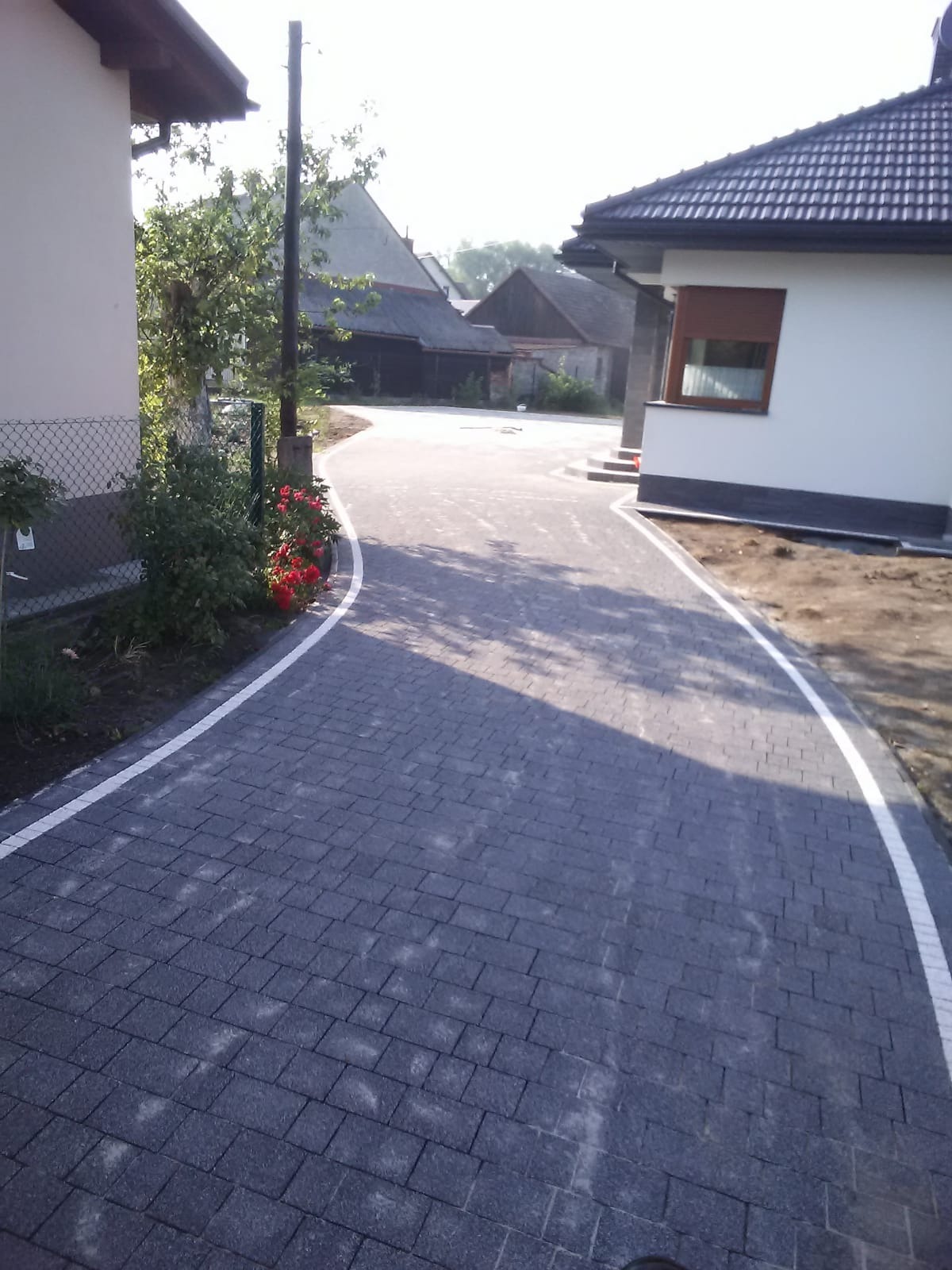 Nowoczesny podjazd z ciemnoszarej kostki brukowej z białymi obrzeżami, prowadzący do domu z brązowymi roletami, z widoczną czerwoną rabatą kwiatową po lewej stronie.