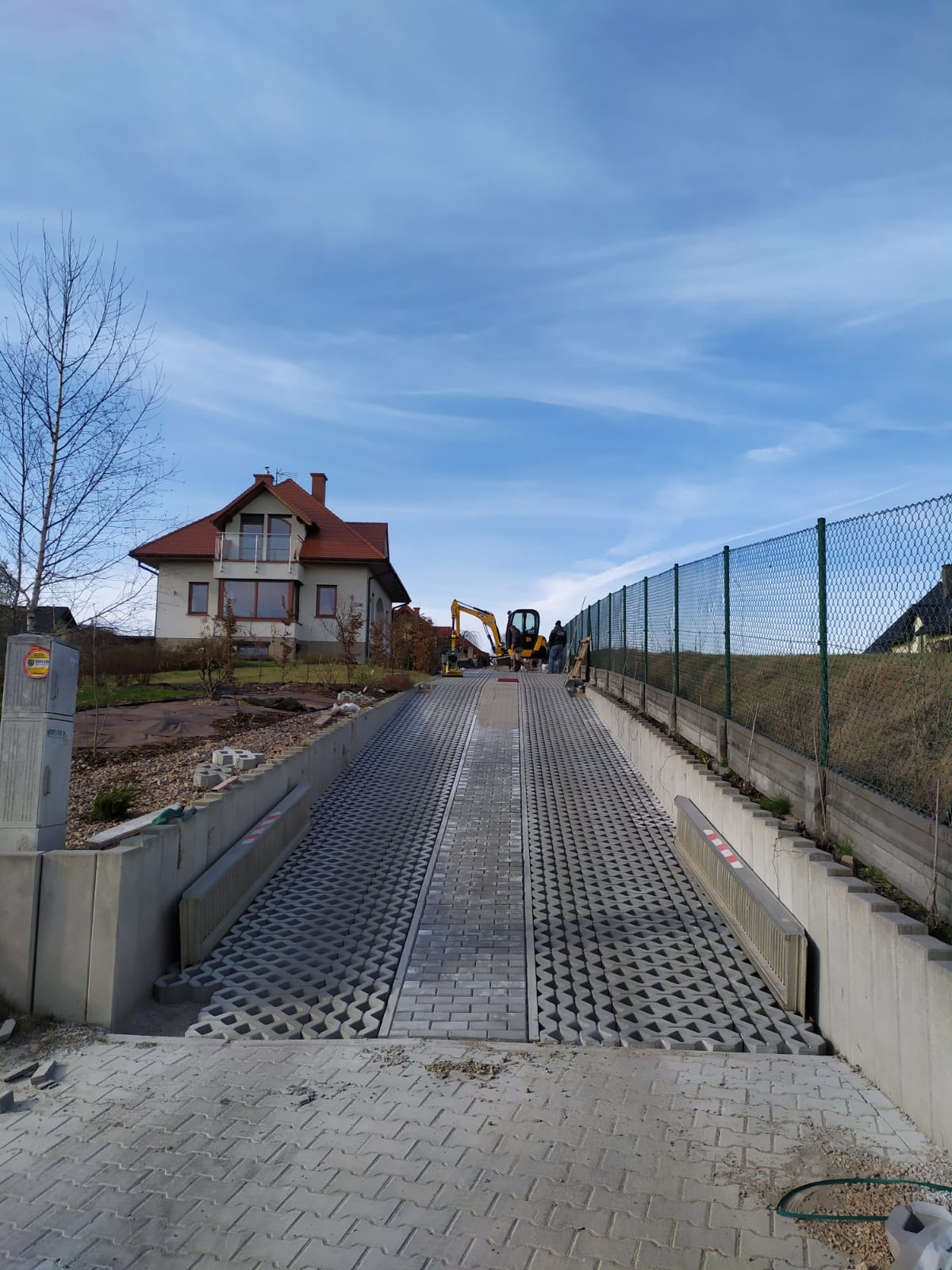 Podjazd z ażurowej kostki brukowej w trakcie budowy, z widocznym minikoparką w tle, otoczony betonowymi krawężnikami i ogrodzeniem z siatki.
