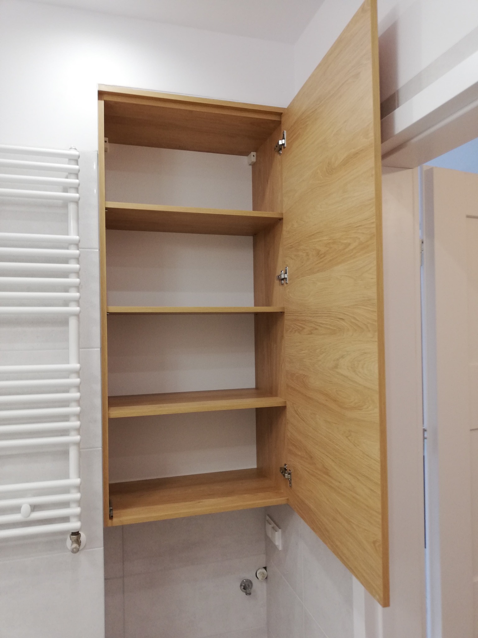 Półka łazienkowa z otwartymi drzwiczkami z jasnego drewna, widoczna na tle białych płytek i grzejnika drabinkowego.