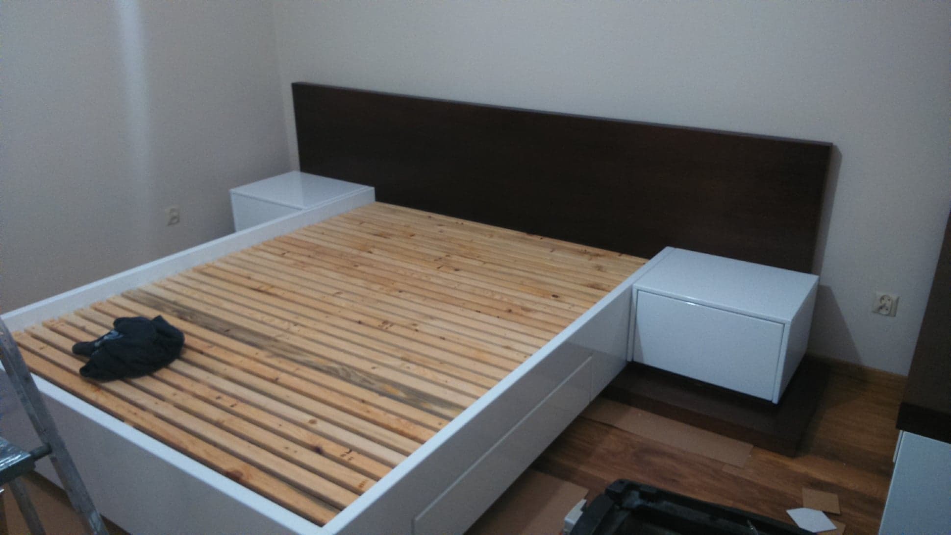 Rama łóżka z białym obramowaniem i drewnianym stelażem, zintegrowana z szafkami nocnymi w kolorze białym i brązowym zagłówkiem, częściowo rozpakowana odzież na stelażu, widoczne narzędzia...