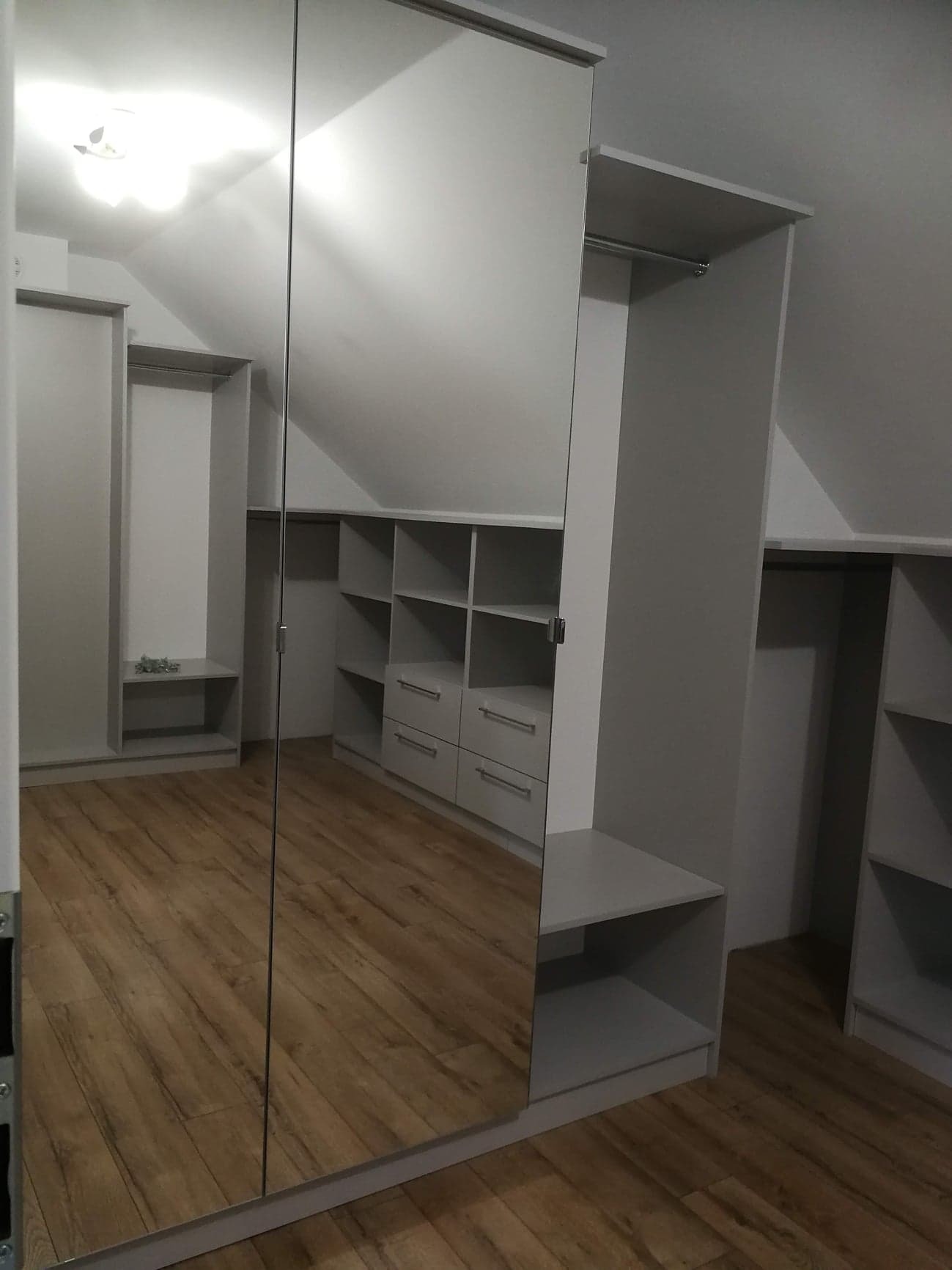 Jasna garderoba na poddaszu ze skośnym sufitem, widoczna przez lustrzane drzwi przesuwne, z półkami, szufladami i drążkami na wieszaki, drewniana podłoga.