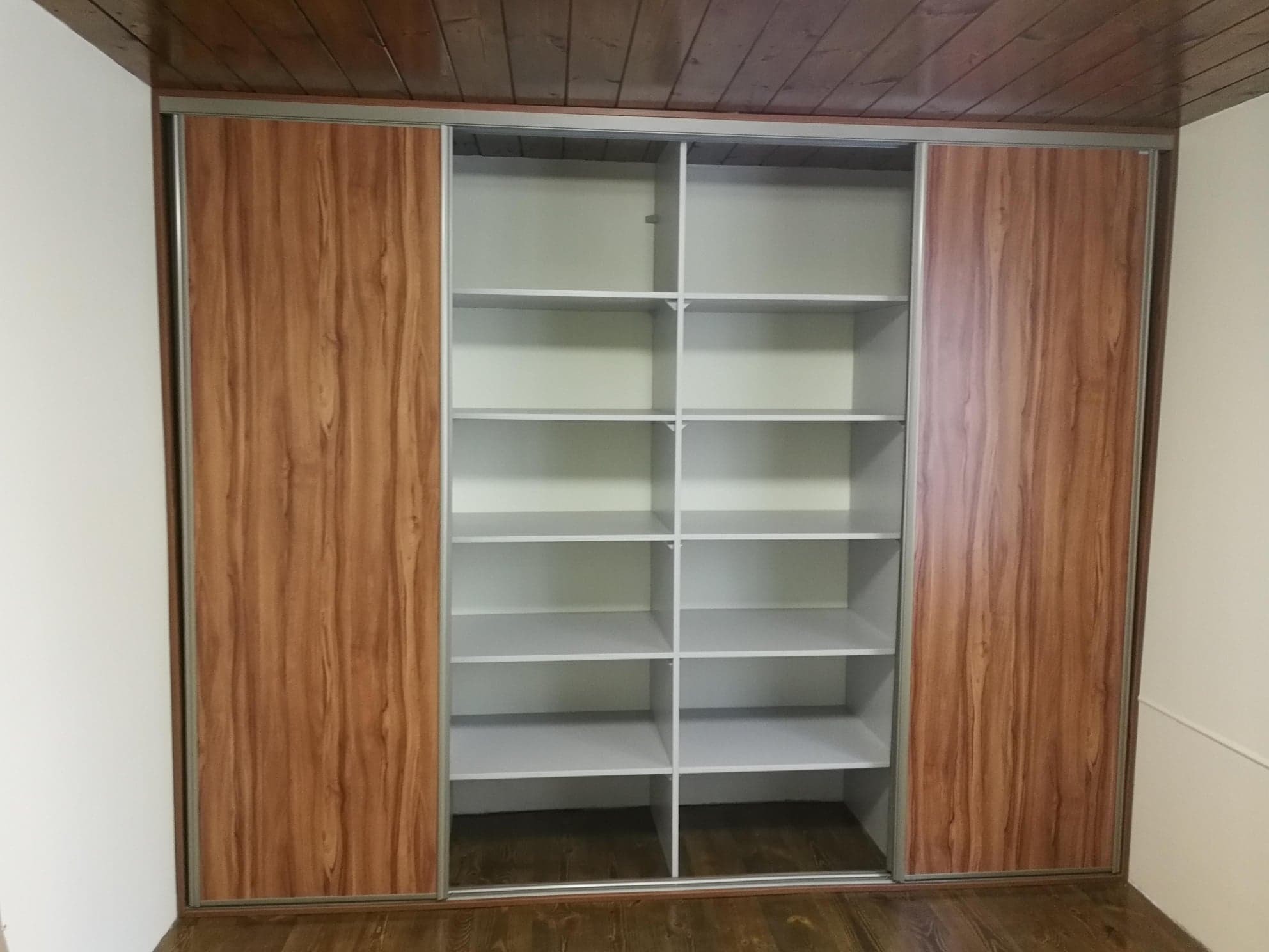 Pusta szafa wnękowa z przesuwnymi drzwiami o wzorze drewna, z widocznymi białymi półkami i drewnianą podłogą oraz sufitem.