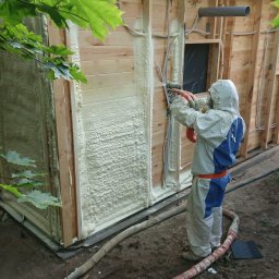 ET Centrum Izolacji - Pracownik w kombinezonie ochronnym aplikuje pianę PUR na drewnianą ścianę budynku, widoczne węże doprowadzające materiał i świeżo nałożona warstwa izolacji.