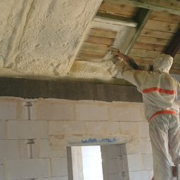 ET Centrum Izolacji - Pracownik w kombinezonie ochronnym, stojący na drabinie, aplikuje pianę PUR na drewnianą konstrukcję poddasza, widoczne ściany z betonu komórkowego.