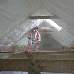 ET Centrum Izolacji - Pracownik w kombinezonie ochronnym aplikuje piankę izolacyjną na poddaszu, widoczne drewniane elementy konstrukcji dachu i fragment instalacji.
