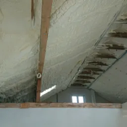 Poddasze ocieplone pianką PUR, widoczne krokwie i instalacje elektryczne, surowe ściany w tle, okno dachowe.