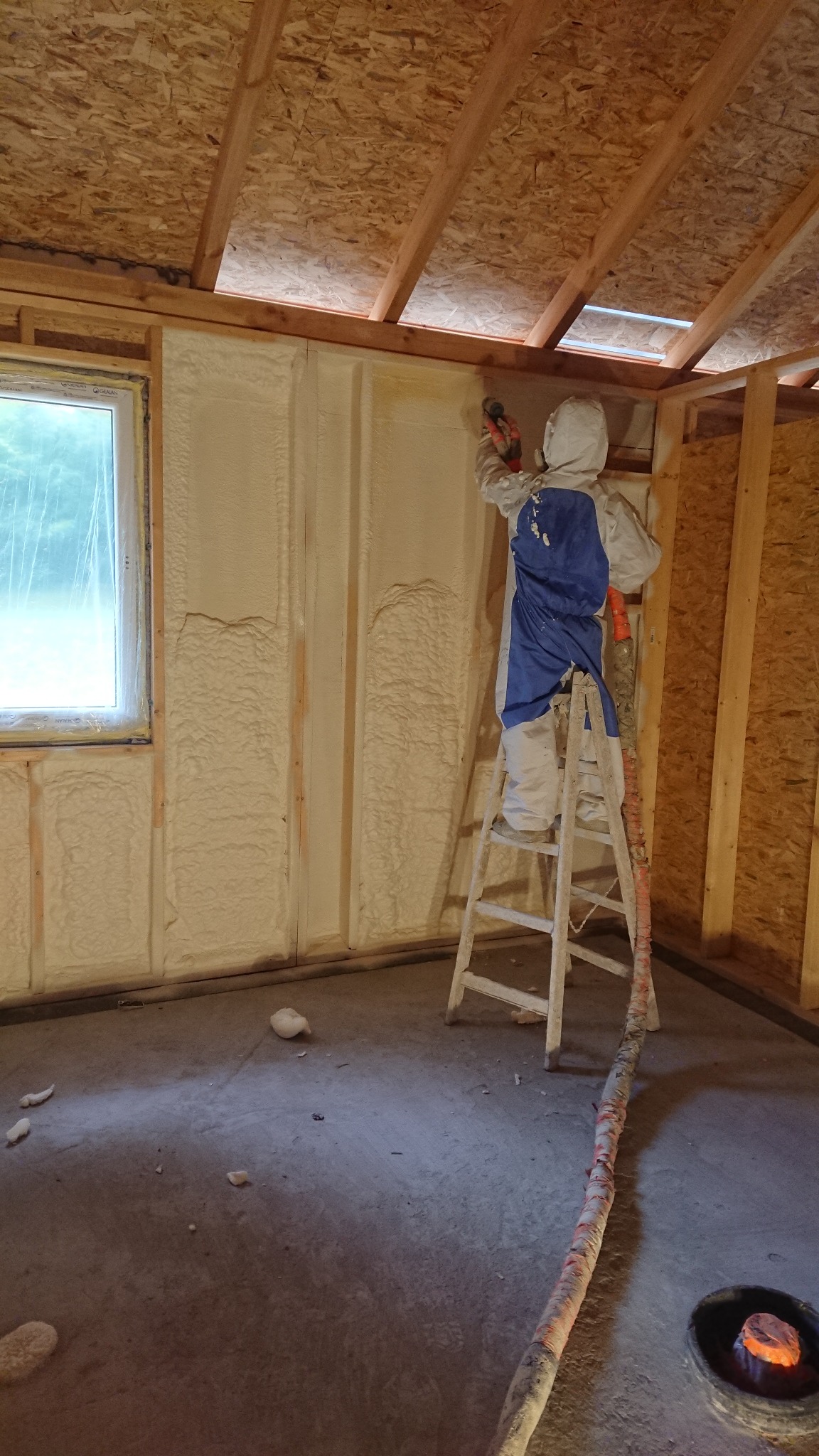 Pracownik w kombinezonie ochronnym aplikuje piankę izolacyjną PUR na ścianę wewnętrzną budynku z drewnianą konstrukcją, stojąc na aluminiowej drabinie. Widoczny wąż doprowadzający piankę...