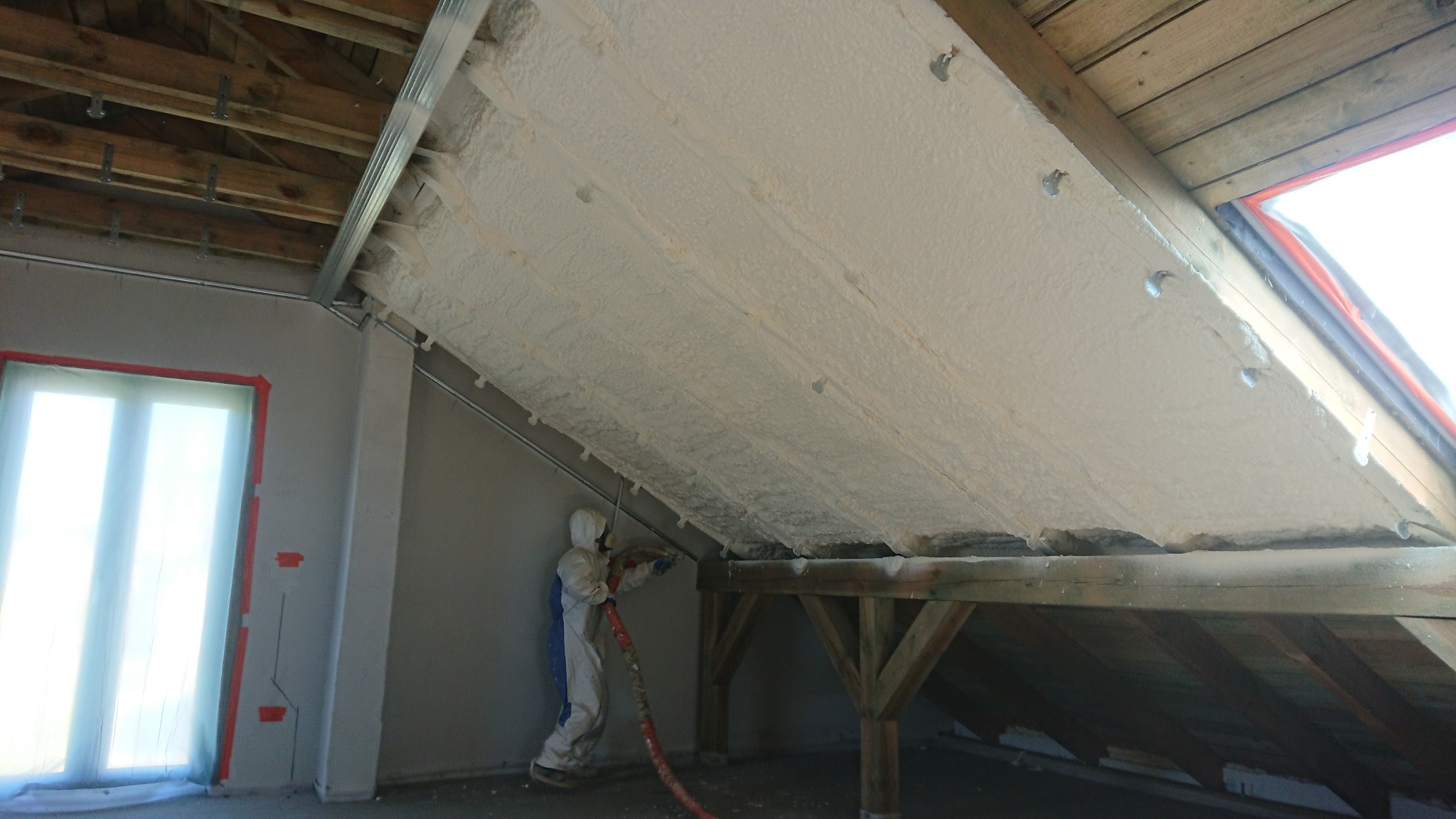 Pracownik w kombinezonie ochronnym aplikuje pianę na poddasze z drewnianą konstrukcją, widoczne metalowe profile mocujące i okno zabezpieczone folią.