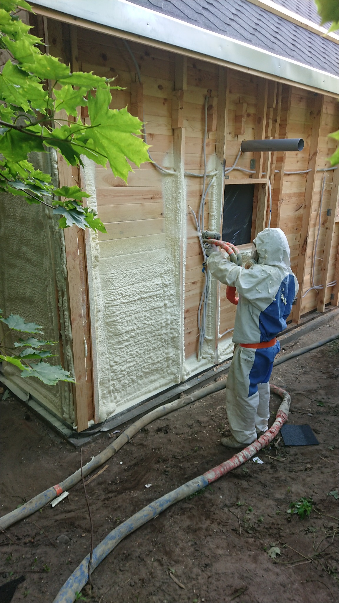 Pracownik w kombinezonie ochronnym aplikuje pianę PUR na drewnianą ścianę budynku, widoczne węże doprowadzające materiał i świeżo nałożona warstwa izolacji.