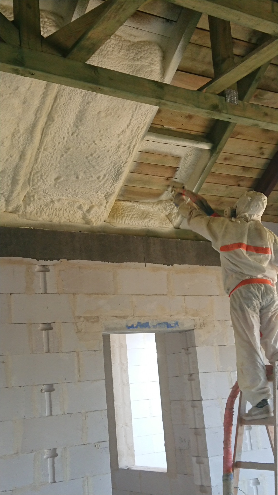 Pracownik w kombinezonie ochronnym, stojący na drabinie, aplikuje pianę PUR na drewnianą konstrukcję poddasza, widoczne ściany z betonu komórkowego.