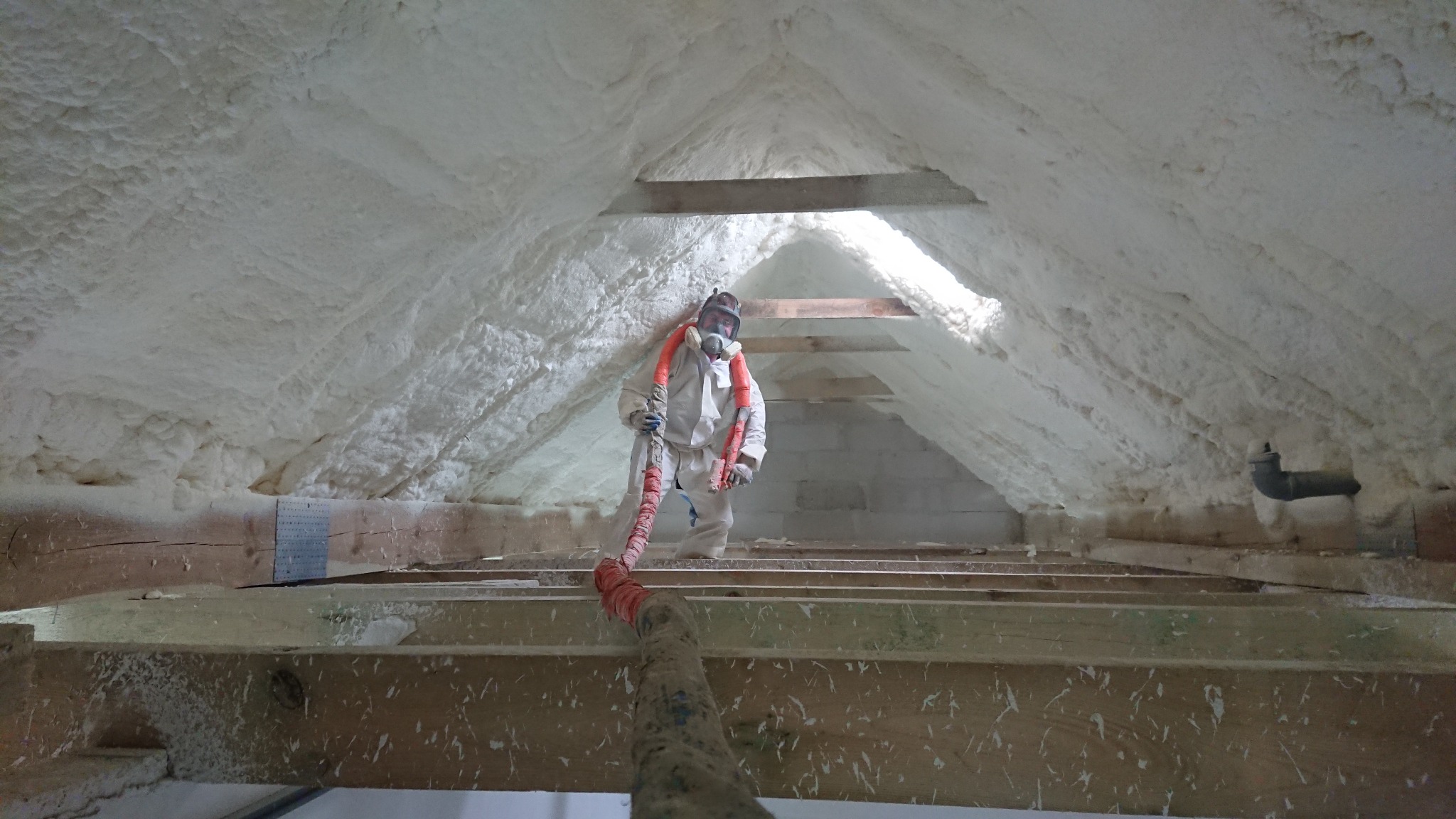 Pracownik w kombinezonie ochronnym aplikuje piankę izolacyjną na poddaszu, widoczne drewniane elementy konstrukcji dachu i fragment instalacji.