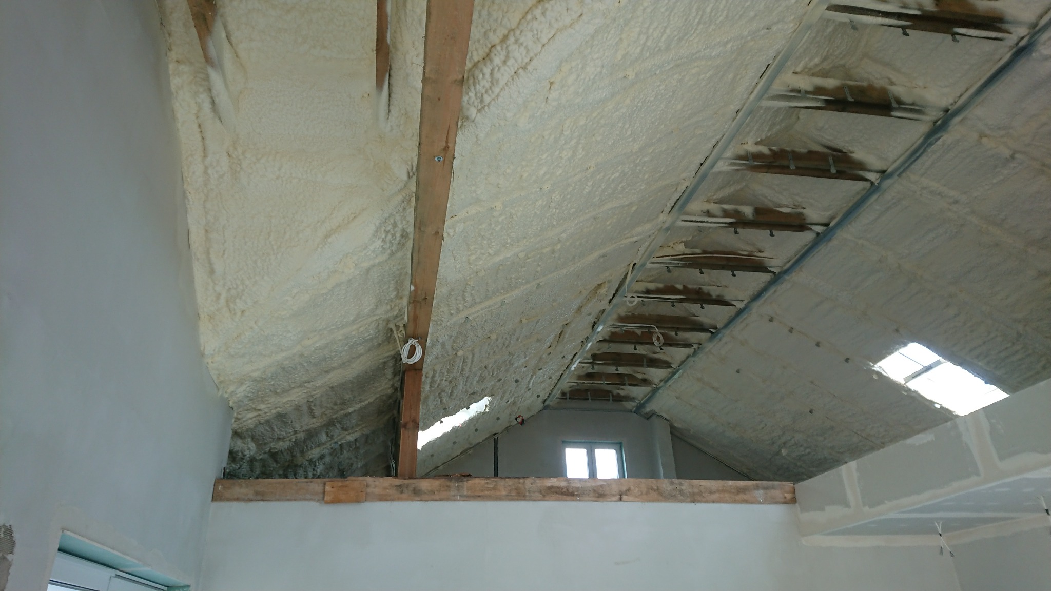 Poddasze ocieplone pianką PUR, widoczne krokwie i instalacje elektryczne, surowe ściany w tle, okno dachowe.