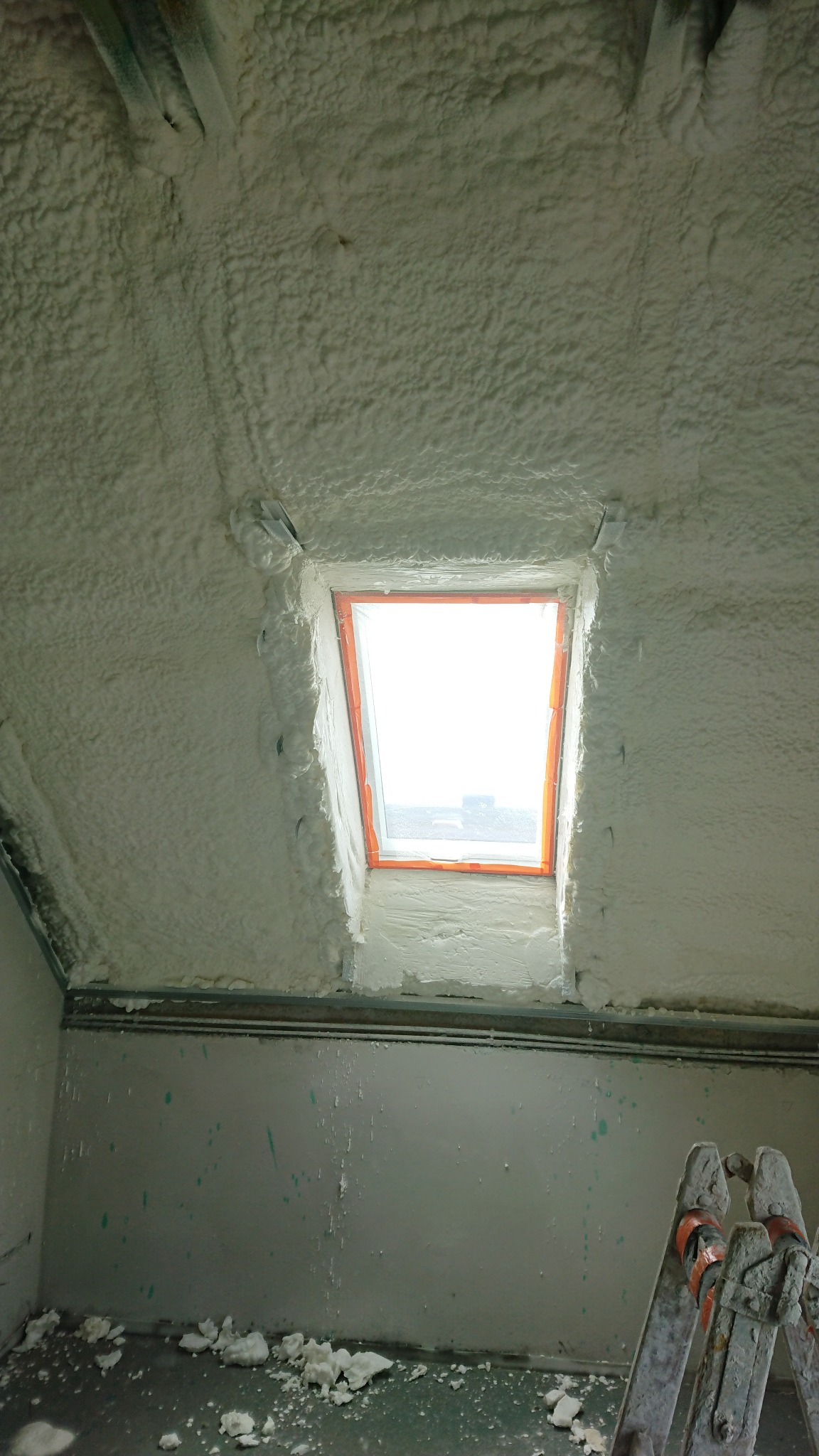 Poddasze po ociepleniu pianką PUR, widoczne okno dachowe w pomarańczowej ramie, fragment drabiny i resztki pianki na podłodze.