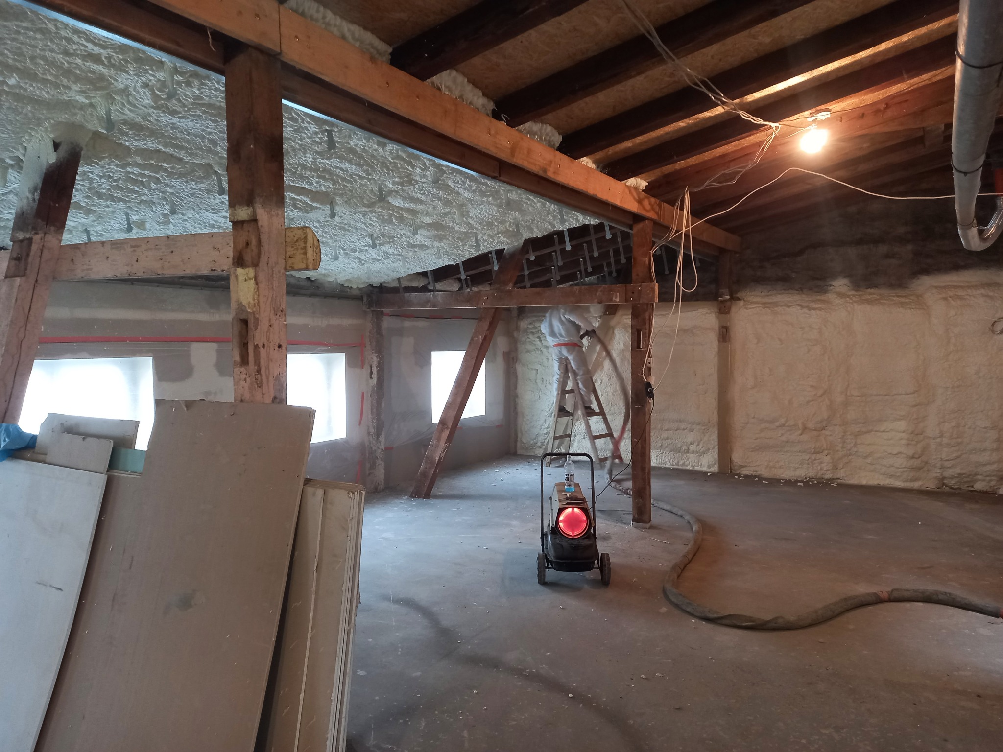 Poddasze ocieplane pianką PUR, widoczny pracownik na drabinie aplikujący pianę, drewniana konstrukcja dachu, nagrzewnica na kółkach, płyty gipsowe w rogu pomieszczenia.