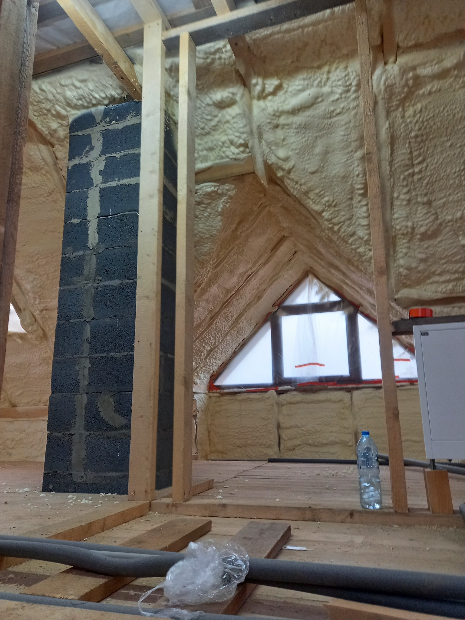 Poddasze ocieplone pianką PUR, widoczny fragment komina z bloczków betonowych, drewniana konstrukcja dachu i okno w szczycie ściany