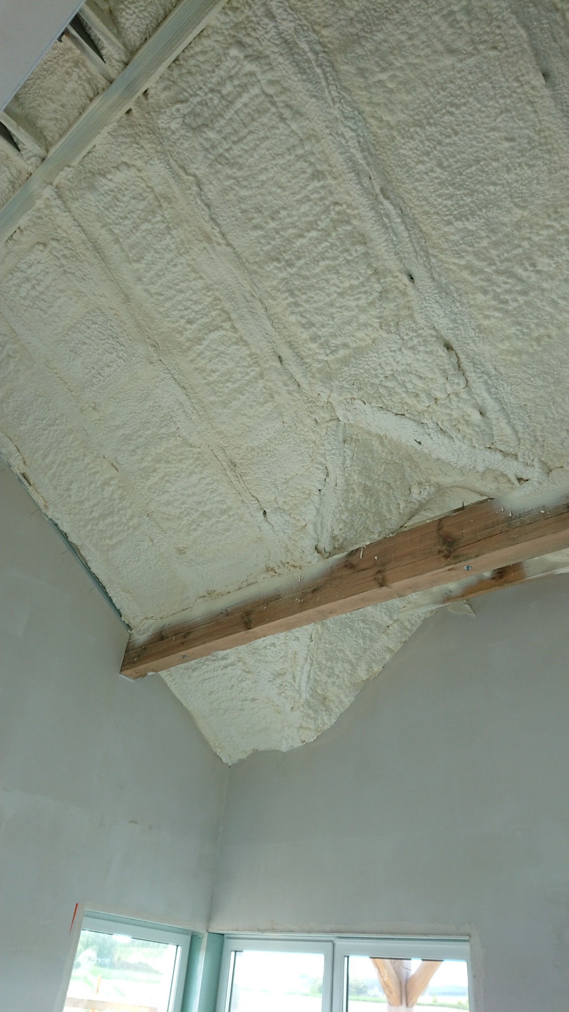 Poddasze z widoczną strukturą pianki izolacyjnej, przytwierdzone drewniane belki, surowe ściany i okno w dolnej części kadru.