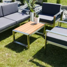 Zestaw mebli ogrodowych: sofa dwuosobowa, dwa fotele i stolik kawowy z drewnianym blatem i metalową ramą, ustawione na trawie. W tle rośliny ozdobne i ogrodzenie.